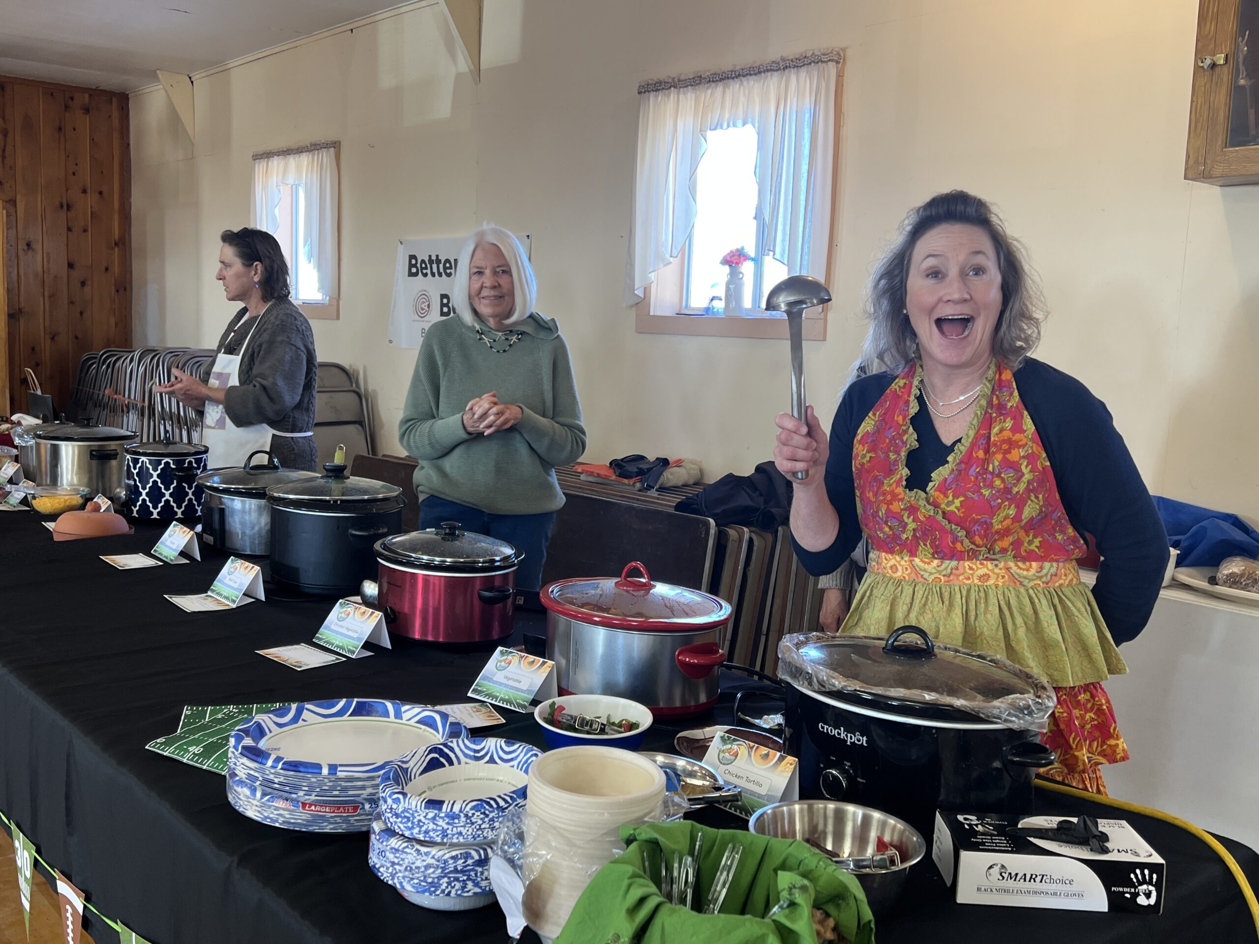 three women behind a table full of food, ready to serve. One woman is holding up a soup ladle.