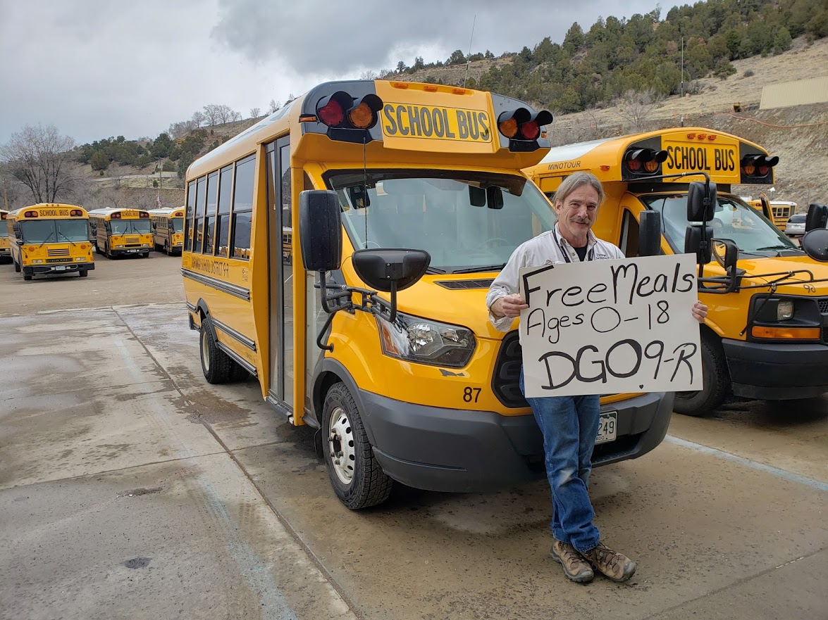 School bus driver, holding 9R Free Meal Pick Up Program sign, for 0-18 yr olds