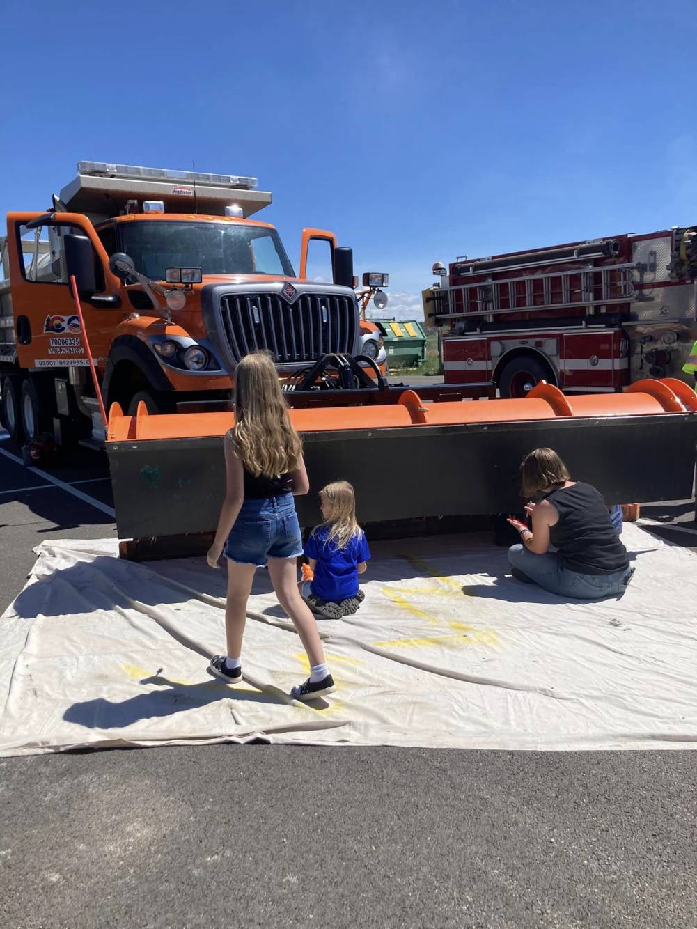 An adult and a child paint iages on the blade of a snowplow while another child looks on. 